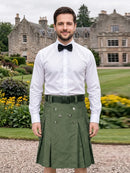 A man wearing a white long-sleeve dress shirt, black bow tie, and an olive green double-breasted kilt stands outdoors in front of a large stone mansion and manicured gardens.