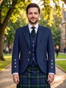 A man wearing a traditional Scottish formal outfit, including a navy blue blazer, matching waistcoat, a green and blue tartan kilt, a white shirt, and a dark tie, standing outdoors on a stone path with trees in the background.