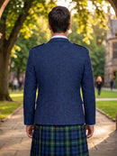 A man viewed from behind wearing a navy blue tweed blazer and a green and blue tartan kilt, standing on a stone path in a sunny park with trees and a historic building in the background.
