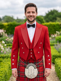 A man wearing a traditional Scottish formal outfit with a bright red jacket and waistcoat, a red tartan kilt, a white shirt, a black bow tie, and a silver sporran, standing in a garden setting.