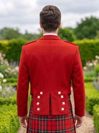 A man viewed from behind wearing a bright red kilt jacket with silver-colored buttons and a red tartan kilt, standing in a garden setting.