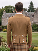 A man viewed from behind wearing a tan Scottish kilt jacket with silver buttons and a tartan kilt, standing in a garden with a stone mansion in the background.